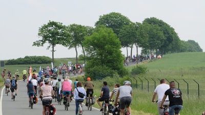 A large group of people is cycling together on a rural road, surrounded by green fields and trees. The cyclists are wearing casual clothing, and some are equipped with helmets. The road is slightly winding and appears to be part of a peaceful countryside setting.