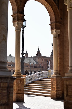 An architectural scene featuring large stone arches and columns framing a view of a historic building with ornate towers. A staircase with intricate railings leads up, while a vintage street lamp is visible. The structure is built of red brick, and the lighting suggests a sunny day.