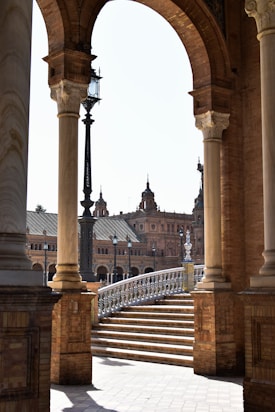 An architectural scene featuring large stone arches and columns framing a view of a historic building with ornate towers. A staircase with intricate railings leads up, while a vintage street lamp is visible. The structure is built of red brick, and the lighting suggests a sunny day.