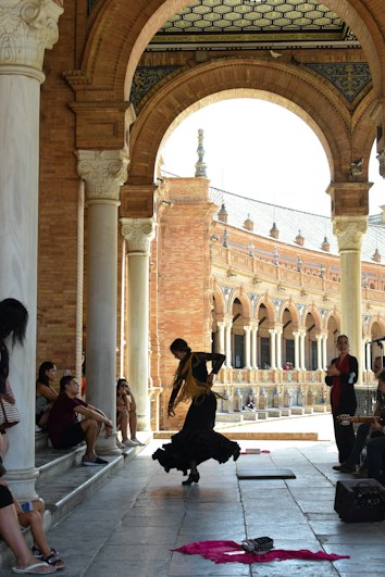 A flamenco dancer performs passionately under an ornate archway, surrounded by a small group of seated spectators. The architecture features intricate brickwork and decorative columns. The dancer wears a traditional outfit, accentuated by a flowing skirt and shawl, while a guitar player accompanies nearby.