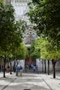 A courtyard lined with lush green trees leads to the entrance of what appears to be a historic or religious building, featuring intricate stone carvings and a decorative facade. Groups of people are gathered around, some taking photographs, suggesting a tourist attraction.