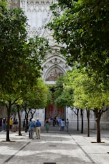 A courtyard lined with lush green trees leads to the entrance of what appears to be a historic or religious building, featuring intricate stone carvings and a decorative facade. Groups of people are gathered around, some taking photographs, suggesting a tourist attraction.