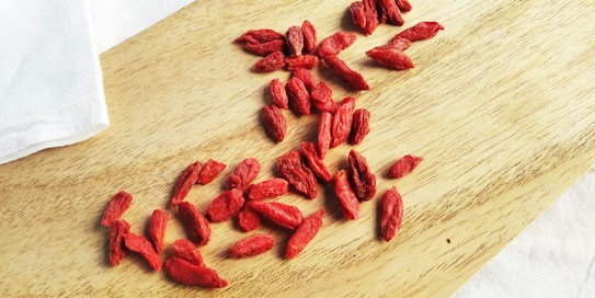 A collection of dried goji berries scattered across a light wooden surface with a white cloth partially visible in the background.