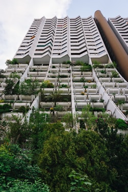 A tall residential building is captured from the ground up, featuring a distinctive zigzag architectural design. The lower levels are covered with abundant greenery, including various shrubs and small trees, giving the structure a seamless blend with nature. The upper floors consist of uniformly designed balconies.