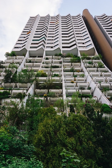 A tall residential building is captured from the ground up, featuring a distinctive zigzag architectural design. The lower levels are covered with abundant greenery, including various shrubs and small trees, giving the structure a seamless blend with nature. The upper floors consist of uniformly designed balconies.