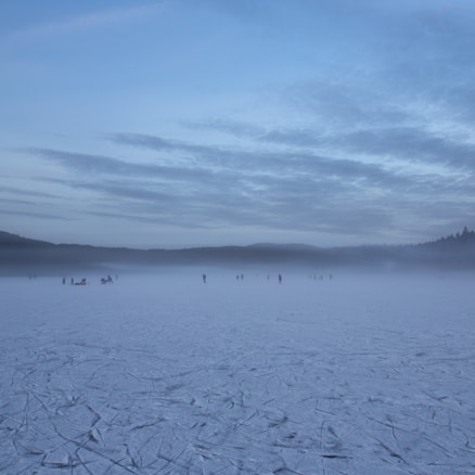 A frozen lake with a misty, overcast sky. Distant figures are walking and engaging in activities on the ice. Trees and hills are faintly visible on the horizon, adding to the serene and cold atmosphere.