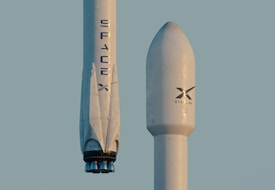 Two white rocket structures featuring the SpaceX logo, with the one on the left displaying rocket engines at the bottom and the one on the right labeled with Starlink. Both positioned vertically in front of a clear sky backdrop.