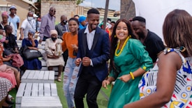 A group of people are gathered outdoors, seemingly enjoying a celebration. Some are seated on white benches, while others are standing or dancing. They are dressed in various colorful outfits, and the mood appears lively and joyful.
