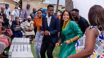 A group of people are gathered outdoors, seemingly enjoying a celebration. Some are seated on white benches, while others are standing or dancing. They are dressed in various colorful outfits, and the mood appears lively and joyful.