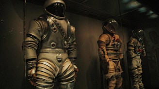 Three vintage space suits are displayed in a dimly lit environment, each suit showcasing a unique design with metallic and fabric components. The suits are positioned upright behind glass, indicating they are part of an exhibit. Shadows and low lighting contribute to a mysterious and historical atmosphere.