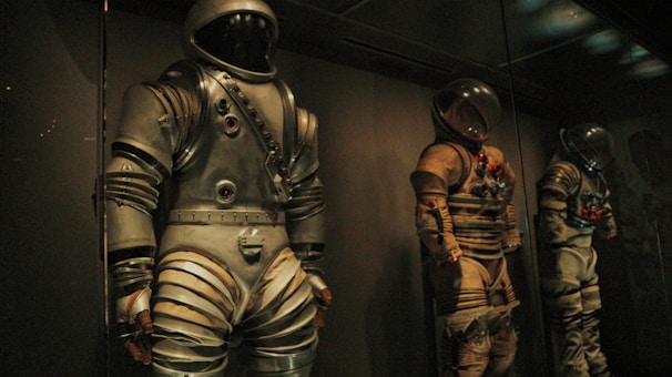 Three vintage space suits are displayed in a dimly lit environment, each suit showcasing a unique design with metallic and fabric components. The suits are positioned upright behind glass, indicating they are part of an exhibit. Shadows and low lighting contribute to a mysterious and historical atmosphere.