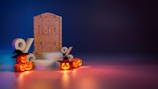 A tombstone with 'RIP' is centered, surrounded by illuminated, carved pumpkins with witch hats. Nearby are large percent symbols and circular platforms, bathed in dramatic orange and blue lighting.