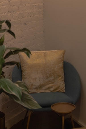 A cozy corner featuring a blue armchair with a large, gold velvet pillow. Next to the chair is a small round wooden side table. A potted plant with long green leaves adds a touch of nature to the space. The background consists of a textured brick wall with a soft, neutral color palette.