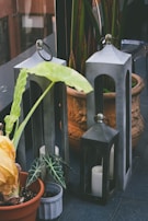 A photograph featuring potted plants and metal lanterns placed on a tiled floor. The plants include a large-leafed green plant in an orange pot and a smaller leafed plant in a small grey pot. The lanterns are of varying heights and contain white candles. In the background, there is a large terracotta pot with intricate designs housing additional green plants.