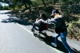 A person in a wheelchair is being pushed by another individual on a pathway surrounded by trees. Sunlight filters through the branches, casting dappled shadows on the ground.