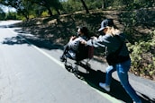 A person in a wheelchair is being pushed by another individual on a pathway surrounded by trees. Sunlight filters through the branches, casting dappled shadows on the ground.