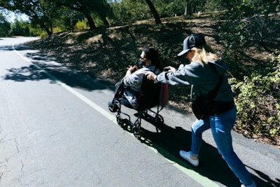 A person in a wheelchair is being pushed by another individual on a pathway surrounded by trees. Sunlight filters through the branches, casting dappled shadows on the ground.