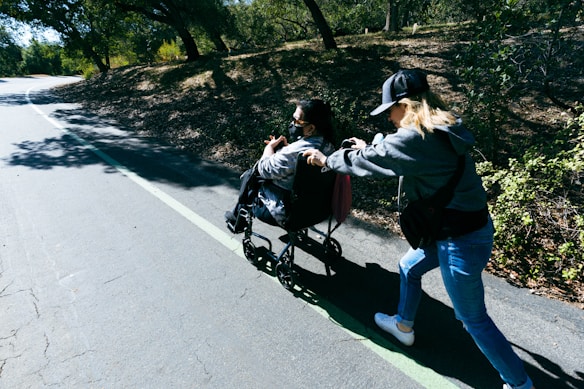 A person in a wheelchair is being pushed by another individual on a pathway surrounded by trees. Sunlight filters through the branches, casting dappled shadows on the ground.