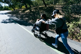 A person in a wheelchair is being pushed by another individual on a pathway surrounded by trees. Sunlight filters through the branches, casting dappled shadows on the ground.