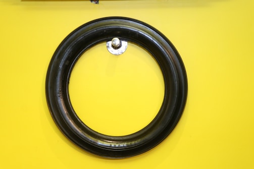 A black circular rubber tire is mounted on a vibrant yellow wall. The tire is centered and appears to be hung by a small metallic hook protruding from the wall.