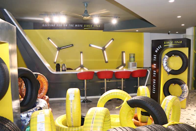 A modern showroom displaying various tires arranged neatly. The background features a bar-like area with red stools and a yellow accent wall. Tire racks display differently wrapped tires against a contrasting black and yellow theme. The lighting is bright, enhancing the contemporary design.