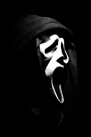A monochrome image of a person wearing a hooded cloak and a white mask with exaggerated features like wide open eyes and mouth. The mask is reminiscent of a classic horror or ghostly visage, with the background obscured in darkness, adding to the spooky and mysterious atmosphere.