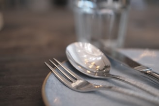 A close-up of a dining table setting featuring a fork, spoon, and knife placed on a light-colored plate. The utensils are shiny and metallic, reflecting light and surroundings. In the background, a glass is visible with a blurred effect, suggesting a shallow depth of field.