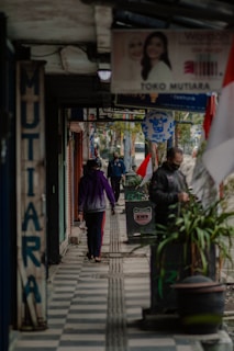 A narrow, covered sidewalk with several pedestrians walking in both directions. Storefront signage and advertisements line the pathway, with a mix of vibrant and muted colors. Several potted plants are placed along the sidewalk, and an Indonesian flag can be seen prominently displayed. The atmosphere is bustling yet routine, with people going about their daily activities.