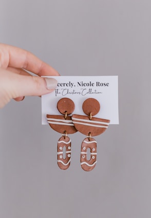 Hand holding a pair of clay earrings attached to a card labeled 'Sincerely, Nicole Rose - The Christmas Collection'. The earrings have rounded and abstract shapes with a brown base color and white decorative lines.