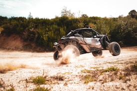 A rugged off-road buggy is kicking up dust as it speeds over a dirt track surrounded by dense greenery. The vehicle appears to be in motion, capturing a dynamic sense of action and adventure.