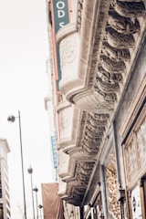 Ornate architectural details on a building facade include intricate carvings and decorative molding. The facade features a repeating pattern of sculpted designs beneath a series of overhangs. Tall street lamps and part of an adjacent building are visible in the background.