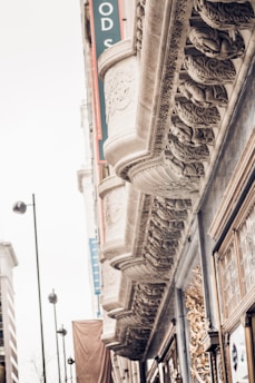 Ornate architectural details on a building facade include intricate carvings and decorative molding. The facade features a repeating pattern of sculpted designs beneath a series of overhangs. Tall street lamps and part of an adjacent building are visible in the background.