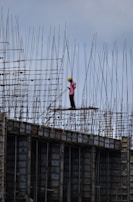 A construction worker wearing a safety helmet stands on a high-rise structure surrounded by a framework of metal rods and beams. The worker appears to be concentrating on a task amidst the towering scaffolding.