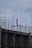 A construction worker wearing a safety helmet stands on a high-rise structure surrounded by a framework of metal rods and beams. The worker appears to be concentrating on a task amidst the towering scaffolding.