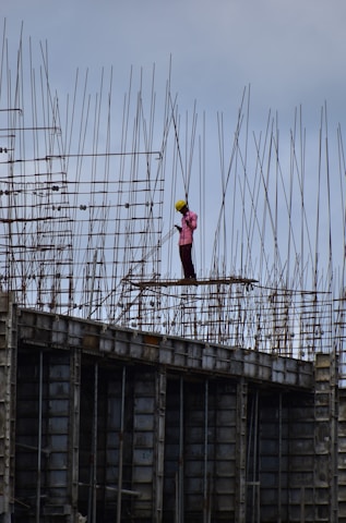 A construction worker wearing a safety helmet stands on a high-rise structure surrounded by a framework of metal rods and beams. The worker appears to be concentrating on a task amidst the towering scaffolding.