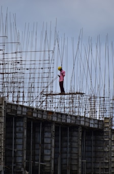A construction worker wearing a safety helmet stands on a high-rise structure surrounded by a framework of metal rods and beams. The worker appears to be concentrating on a task amidst the towering scaffolding.