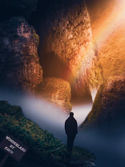 A person stands on a grassy edge overlooking a mystical landscape with steep cliffs and a rising sun casting warm, golden light across the scene. Mist rolls through the valleys, creating a dreamlike atmosphere, and a flock of birds flies through the air. A small wooden sign reads 'Wonderland by Emon.'