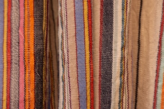 Colorful woven textiles with vertical stripes in various shades including blue, red, yellow, orange, black, and beige. The fabric appears to have a textured surface, typical of traditional woven materials.