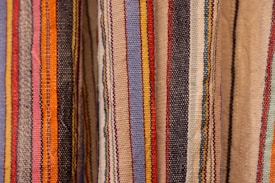 Colorful woven textiles with vertical stripes in various shades including blue, red, yellow, orange, black, and beige. The fabric appears to have a textured surface, typical of traditional woven materials.