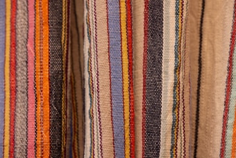Colorful woven textiles with vertical stripes in various shades including blue, red, yellow, orange, black, and beige. The fabric appears to have a textured surface, typical of traditional woven materials.