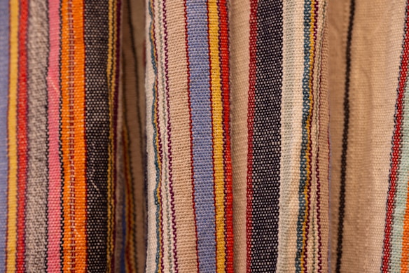 Colorful woven textiles with vertical stripes in various shades including blue, red, yellow, orange, black, and beige. The fabric appears to have a textured surface, typical of traditional woven materials.