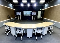 A modern conference room equipped with advanced technology, featuring a curved conference table with five ergonomic office chairs. The room has two large screens at the front displaying video conferencing and presentations. There are cameras set up around the room, likely for video capture or streaming. The walls have a wood panel design, and the lighting is bright and evenly distributed across the space.