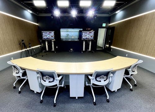 A modern conference room equipped with advanced technology, featuring a curved conference table with five ergonomic office chairs. The room has two large screens at the front displaying video conferencing and presentations. There are cameras set up around the room, likely for video capture or streaming. The walls have a wood panel design, and the lighting is bright and evenly distributed across the space.
