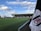 A football stadium with a neatly maintained green field, surrounded by seating stands. The stand in the background prominently displays the word 'FULHAM'. A crane and some construction work are visible on the left, with a partly cloudy sky above. A white corner flag with a red and black logo is prominently visible in the foreground.