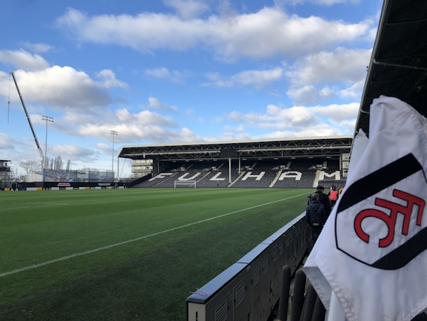 A football stadium with a neatly maintained green field, surrounded by seating stands. The stand in the background prominently displays the word 'FULHAM'. A crane and some construction work are visible on the left, with a partly cloudy sky above. A white corner flag with a red and black logo is prominently visible in the foreground.