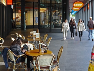 A street scene with an outdoor cafe, several empty tables with woven chairs, and a few people walking past. One person is seated at a table, seemingly engaged with their phone or tablet. The background includes a glass-fronted store with visible signage, and there’s a Blokker store sign visible.