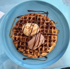 A large, round waffle sits on a blue plate, topped with two scoops of ice cream—one vanilla and one chocolate. The waffle is drizzled with chocolate and caramel sauce, creating a rich, indulgent look.