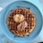 A large, round waffle sits on a blue plate, topped with two scoops of ice cream—one vanilla and one chocolate. The waffle is drizzled with chocolate and caramel sauce, creating a rich, indulgent look.