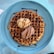 A large, round waffle sits on a blue plate, topped with two scoops of ice cream—one vanilla and one chocolate. The waffle is drizzled with chocolate and caramel sauce, creating a rich, indulgent look.