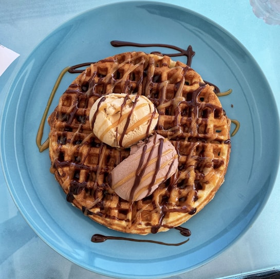 A large, round waffle sits on a blue plate, topped with two scoops of ice cream—one vanilla and one chocolate. The waffle is drizzled with chocolate and caramel sauce, creating a rich, indulgent look.